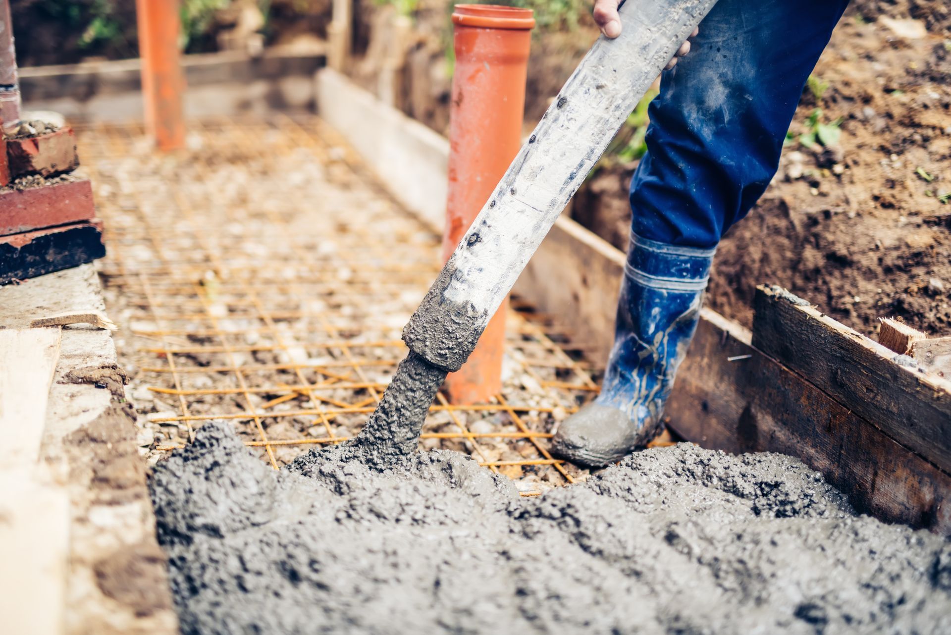 Worker pouring wet concrete at a construction site, with boots, rebar, and orange pipes visible.