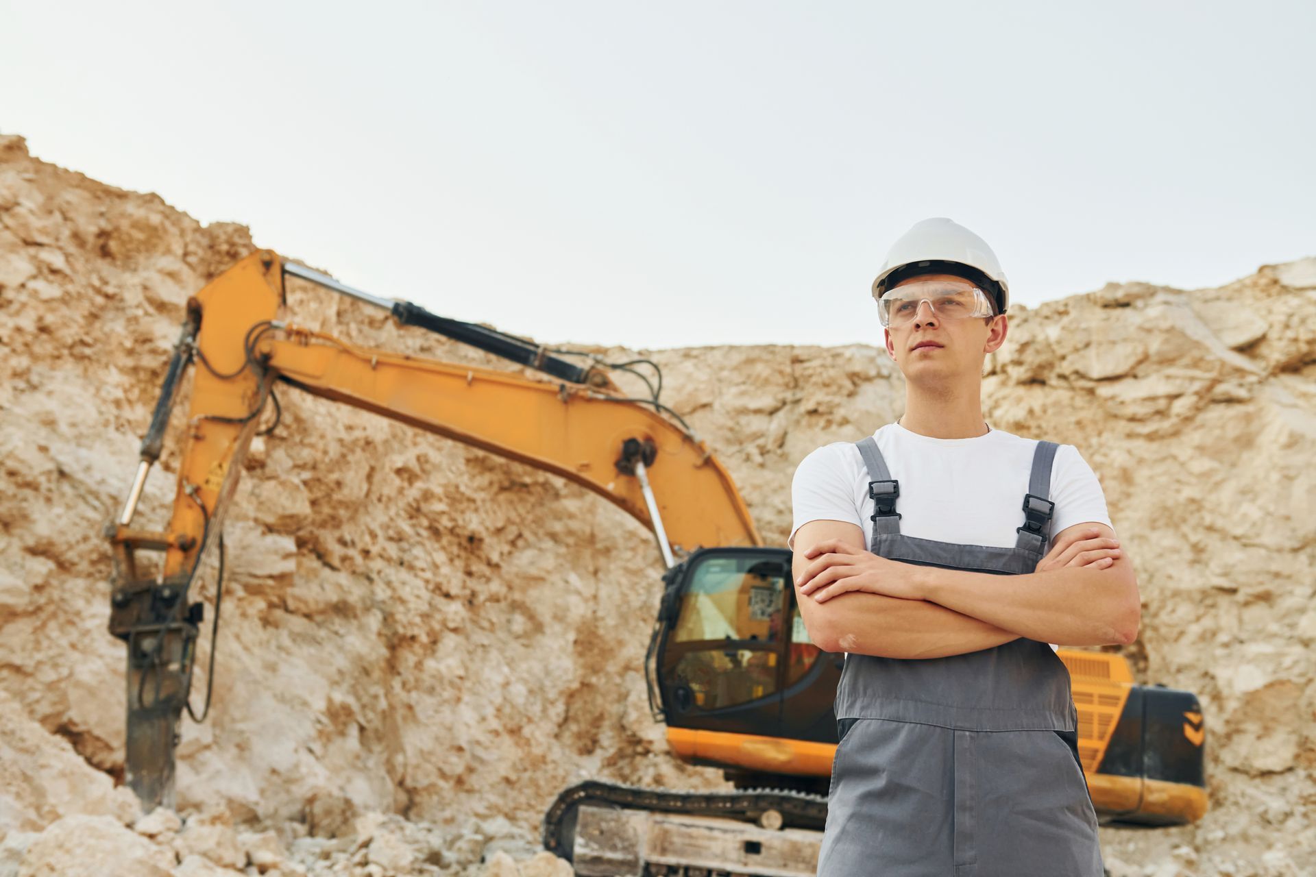 Construction worker standing arms crossed in front of an orange excavator and rocky cliff.