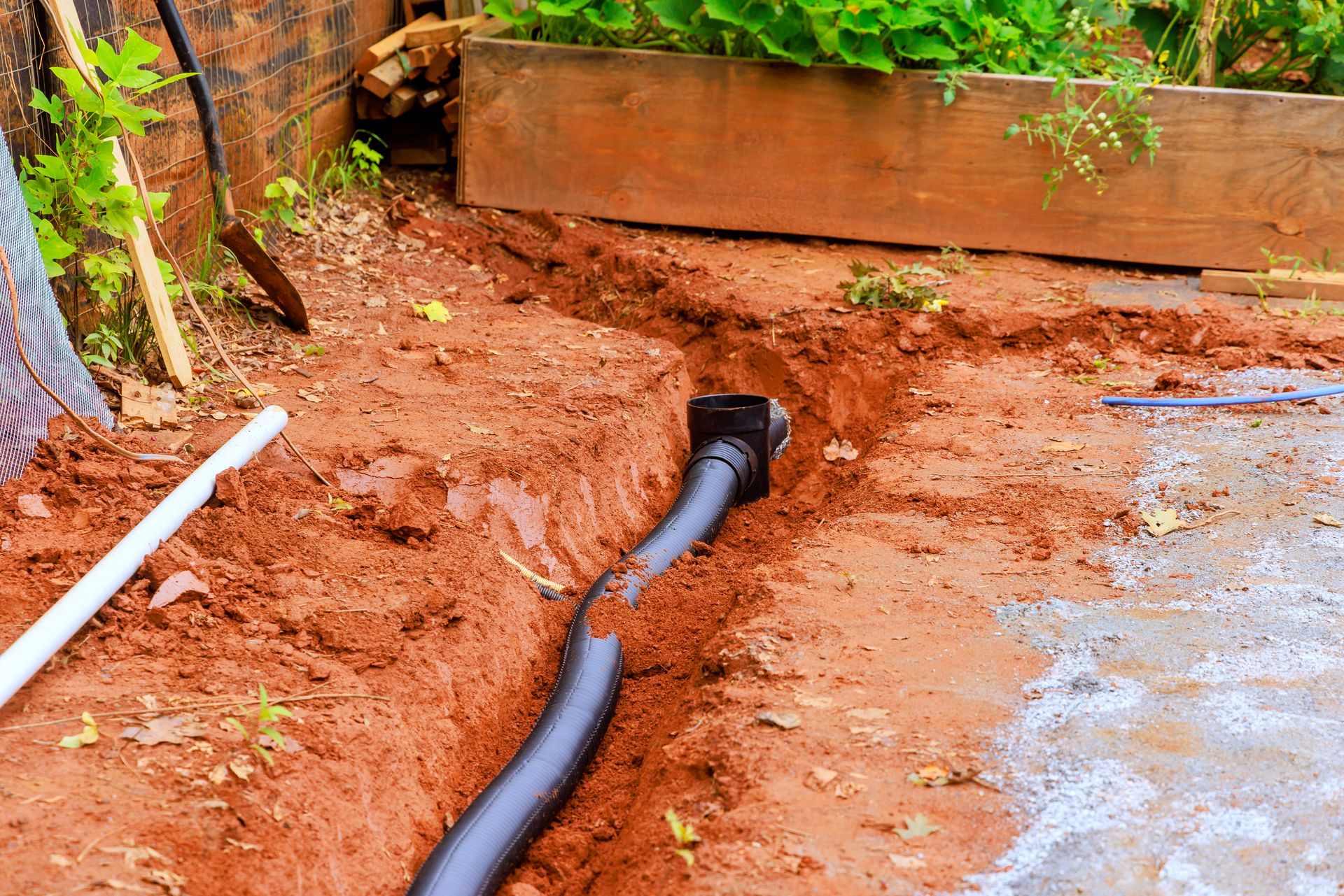 Black pipe laid in a muddy trench beside a brick wall and wooden planter
