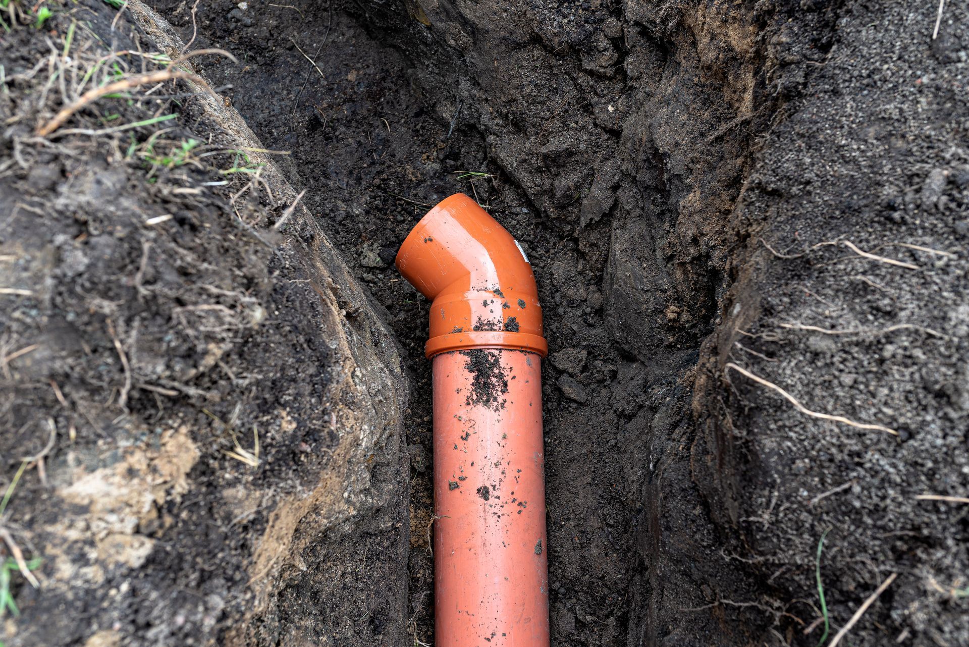 Orange drain pipe elbow in a dirt trench beside dark soil and roots