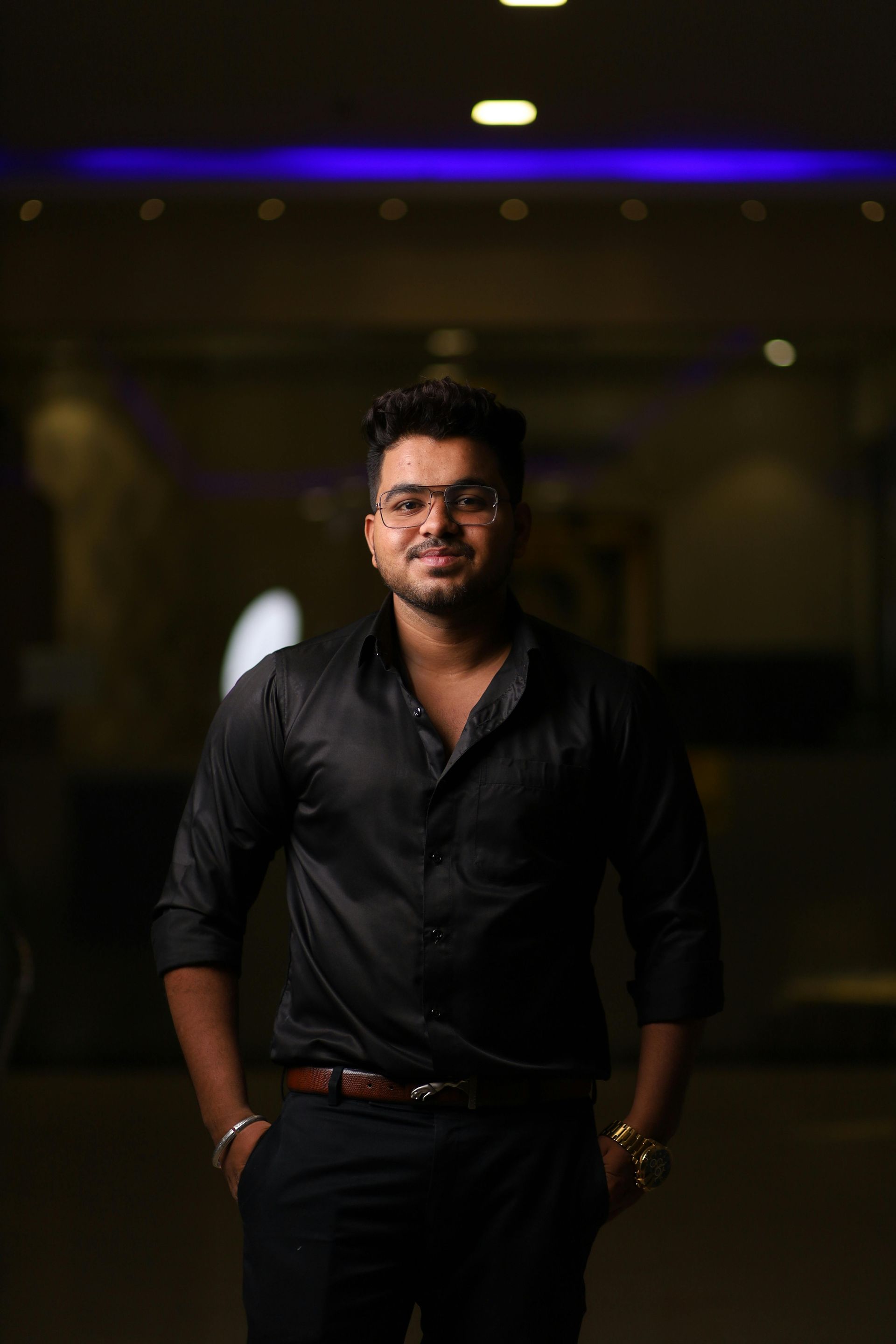 Man in black shirt and pants standing indoors against a dark, softly lit background.