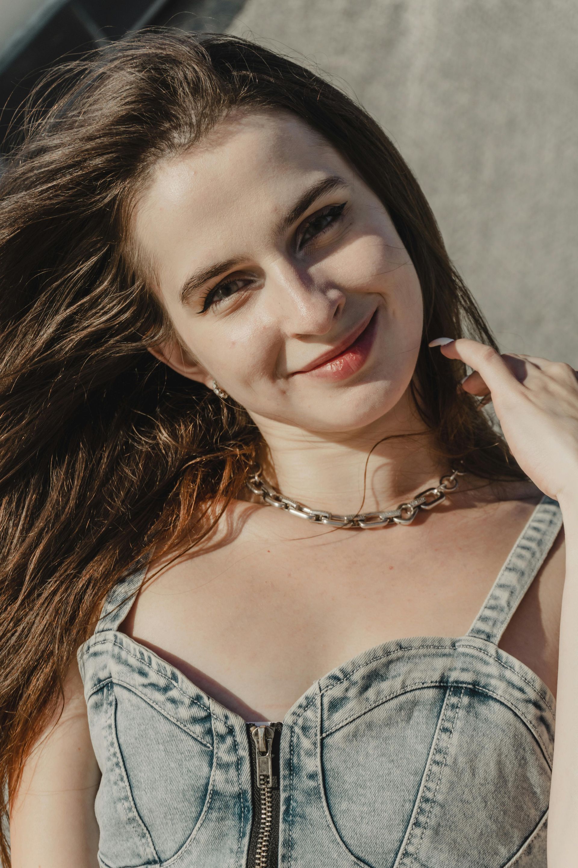 Smiling person with long dark hair in a light denim top, posing outdoors in sunlight.