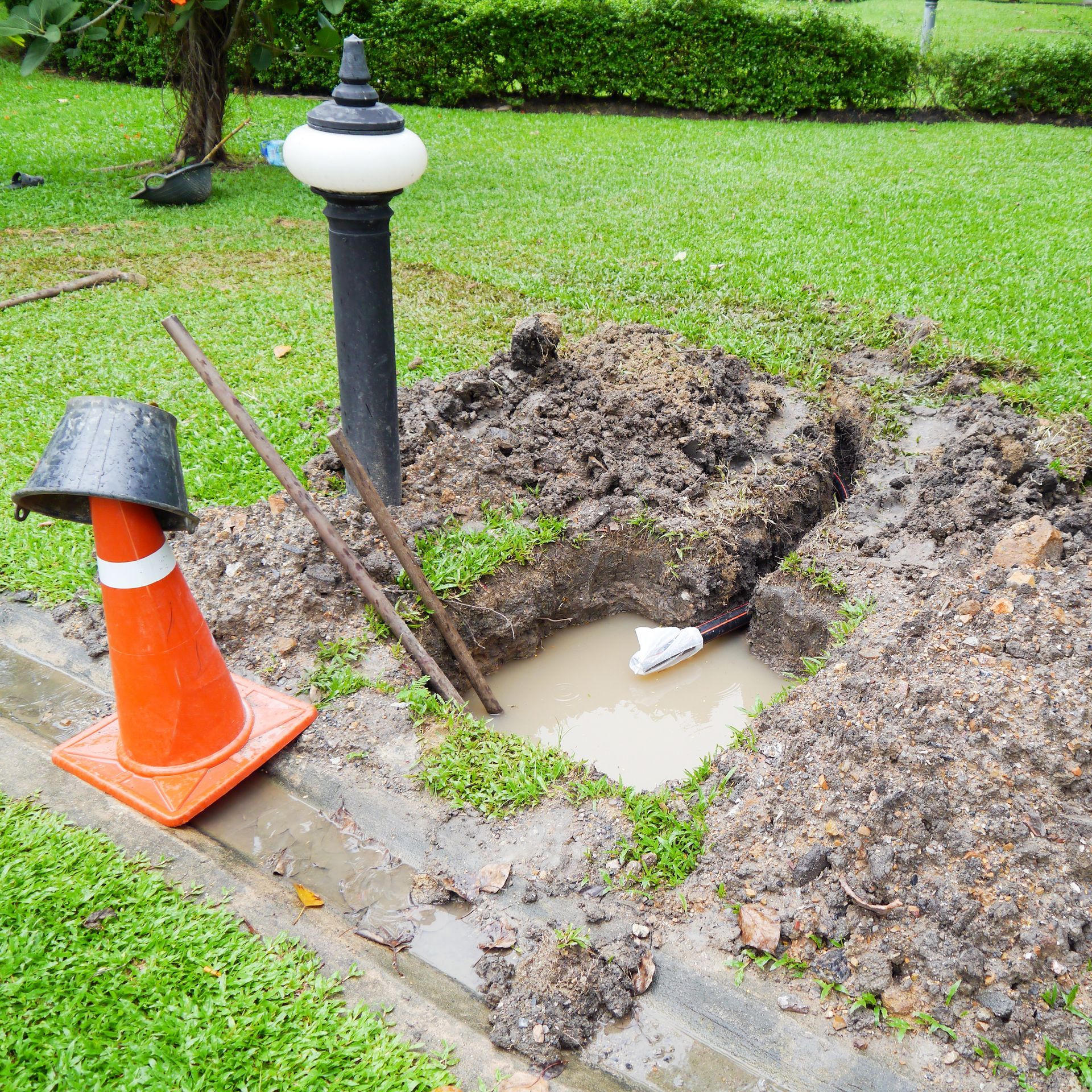 Orange cone and a muddy excavation around a lamp post in a grassy park, with standing water in the pit