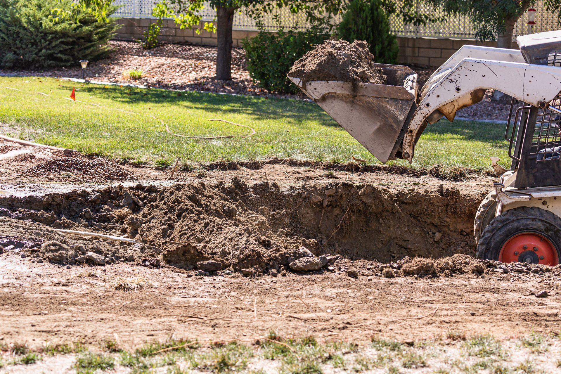 Excavator scooping dirt beside a dug trench in a grassy yard under construction