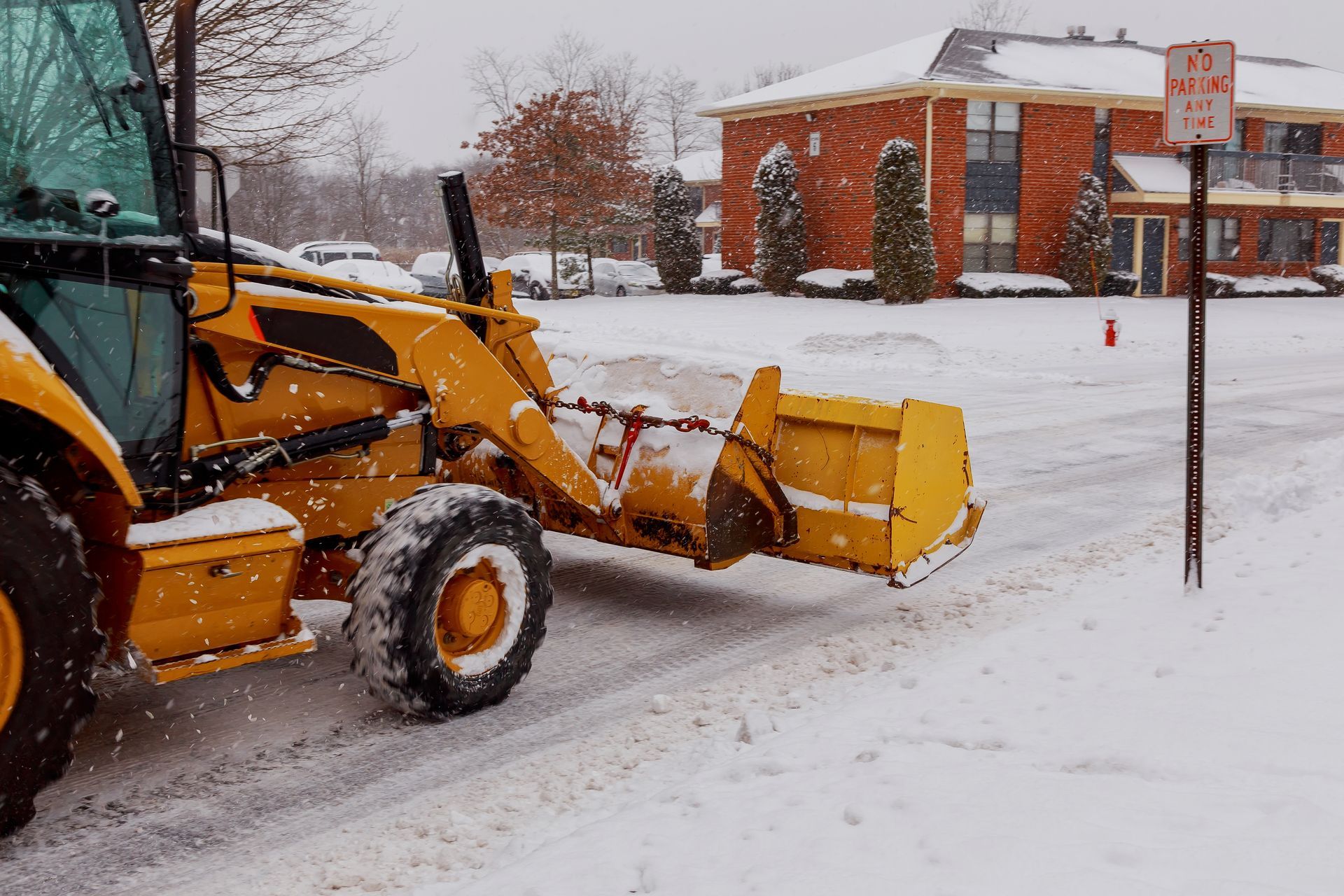 Yellow snowplow clearing a snowy street near a brick building and parking lot