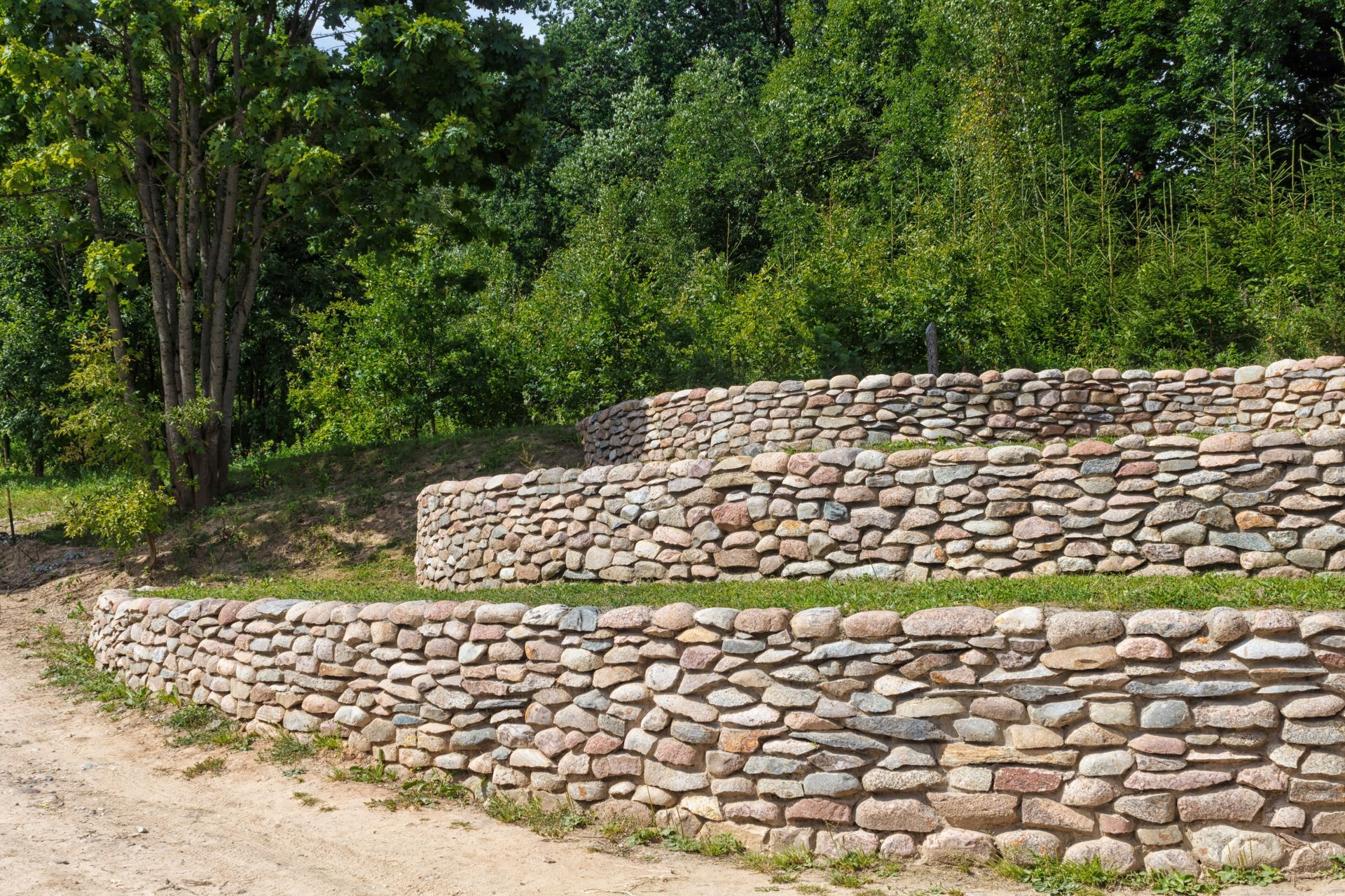 Stone retaining walls beside a dirt path, surrounded by dense green trees and shrubs.