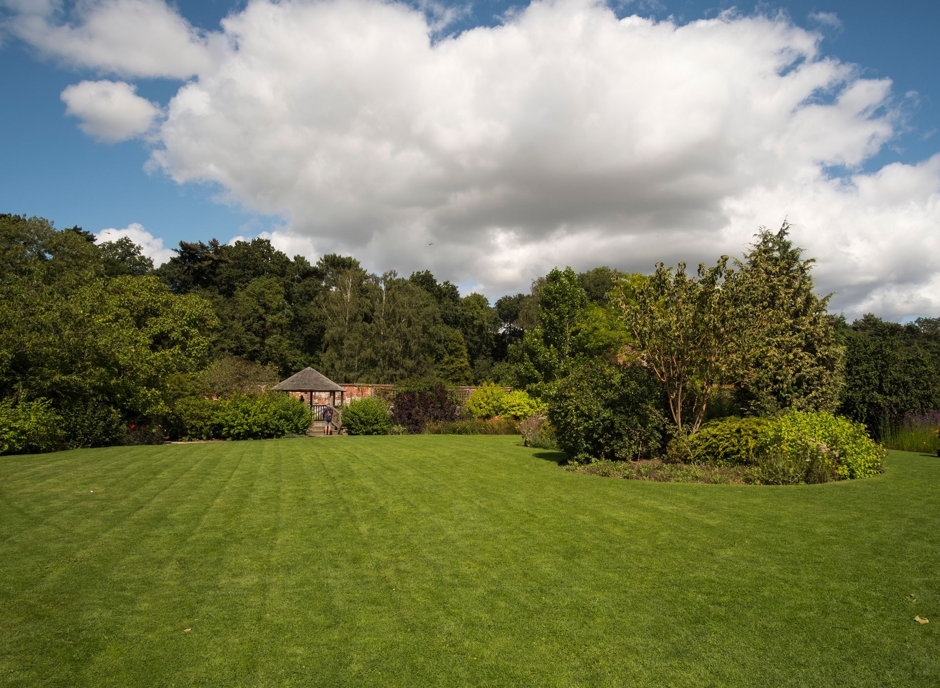 Lush green lawn with trees, shrubs, a small gazebo, and large white clouds overhead