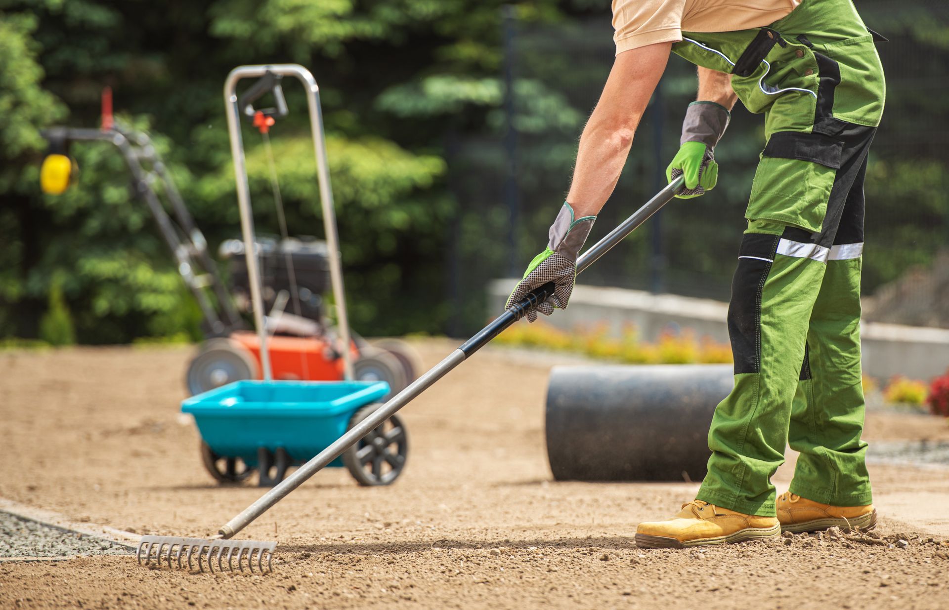 Worker raking gravel on a driveway, with a wheelbarrow and tools in the background