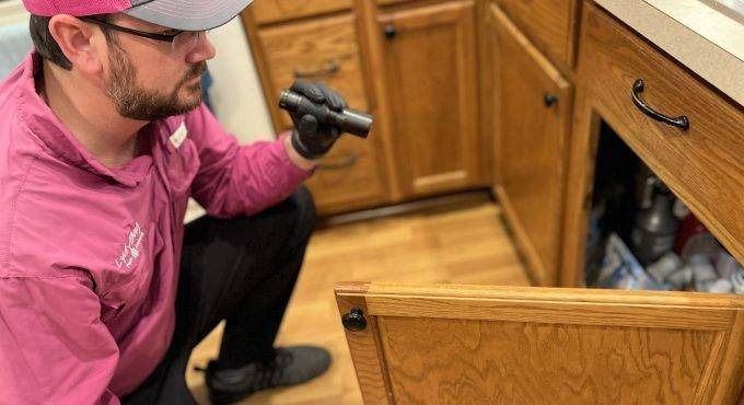 Worker inspecting an open wooden cabinet under a kitchen counter with a flashlight