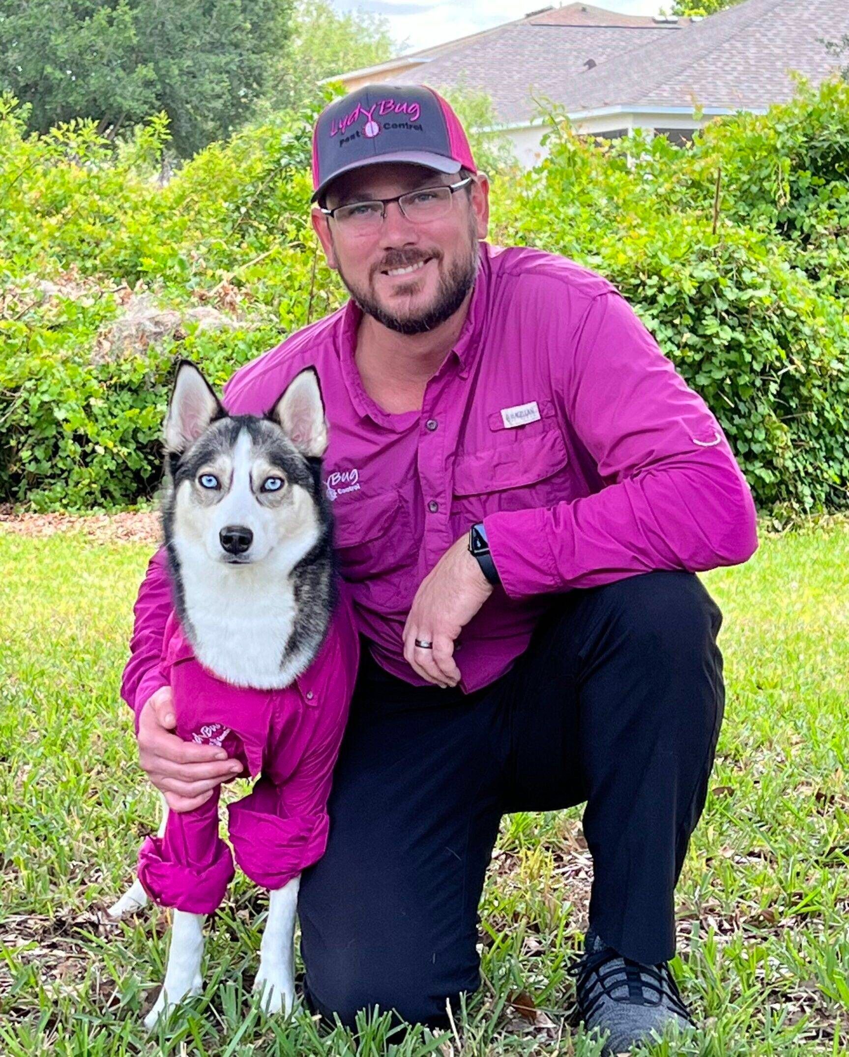 Man kneels beside a husky in a purple jacket in a grassy yard, both wearing matching purple