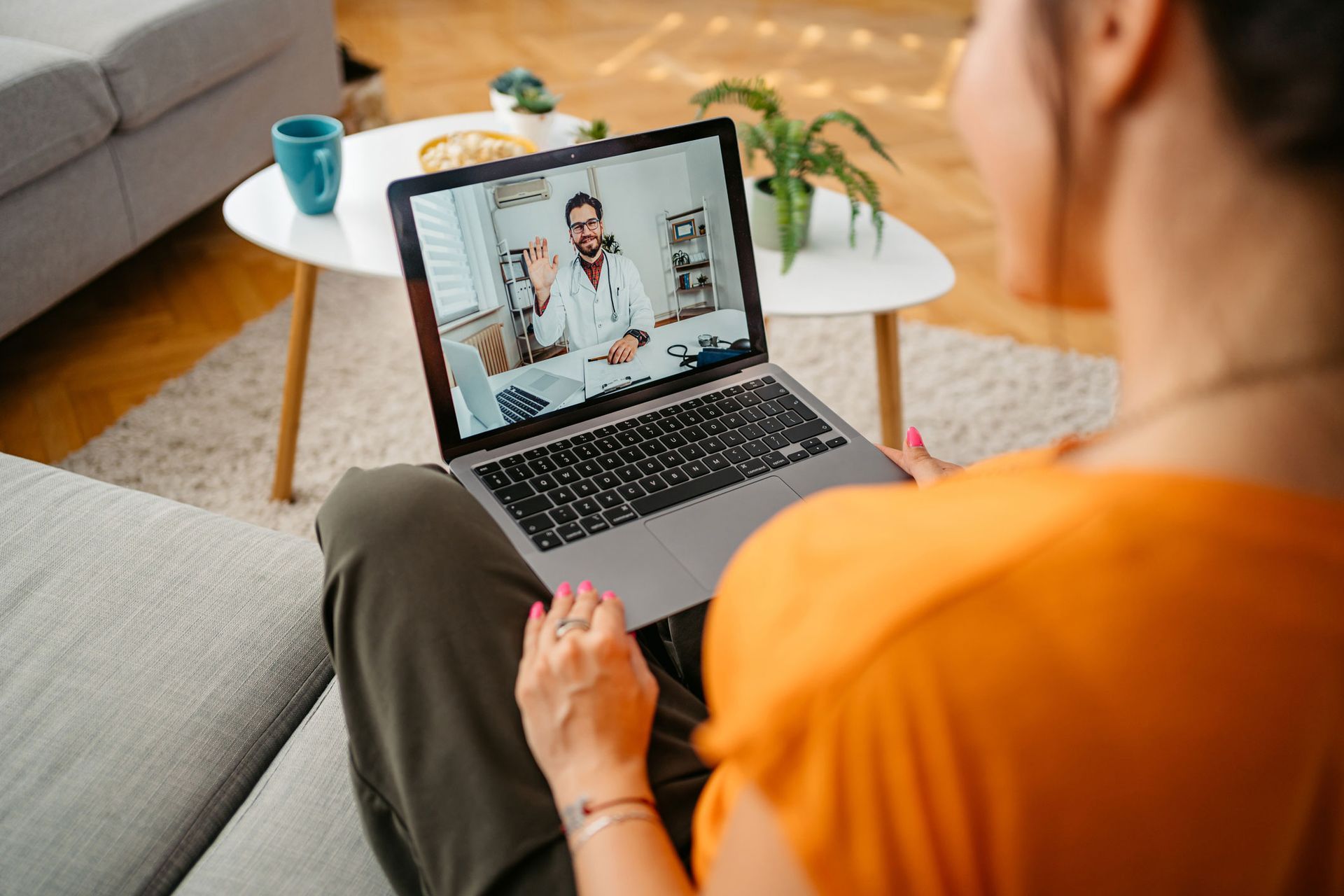 A woman is sitting on a couch using a laptop computer to have a video call with a doctor.