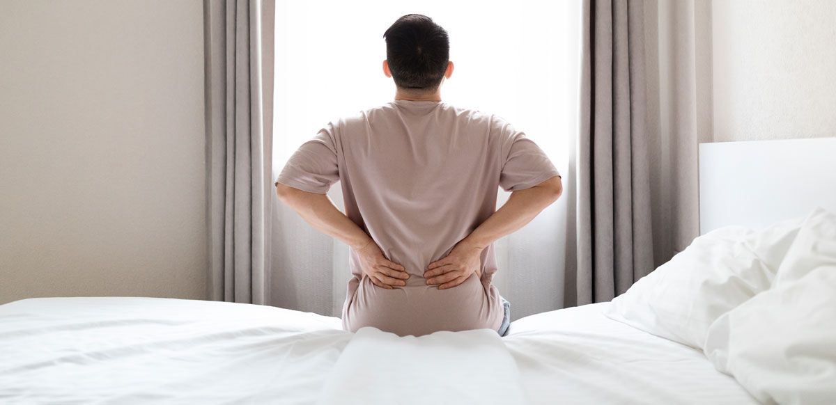Person sitting on a bed, holding their lower back near a window, likely in pain.