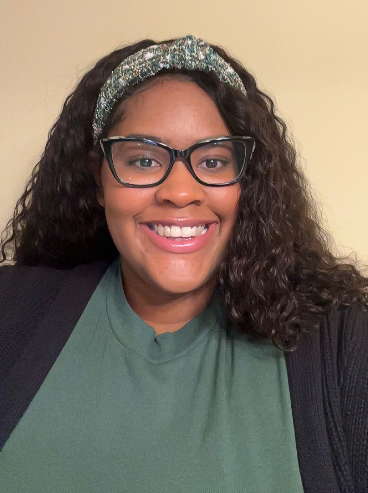 Smiling Black woman with curly hair, glasses, and a headband, wearing a green shirt and black cardigan.