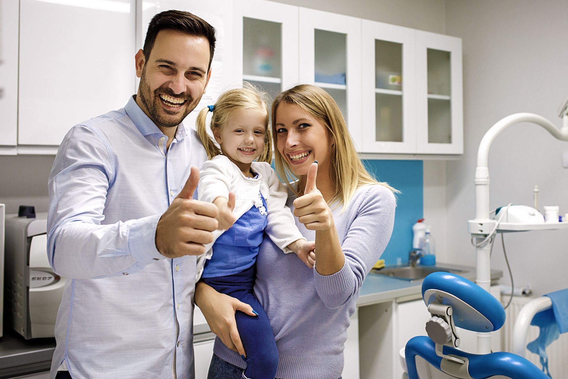 Two adults and a child giving thumbs up inside a dental clinic, with a dental chair visible.