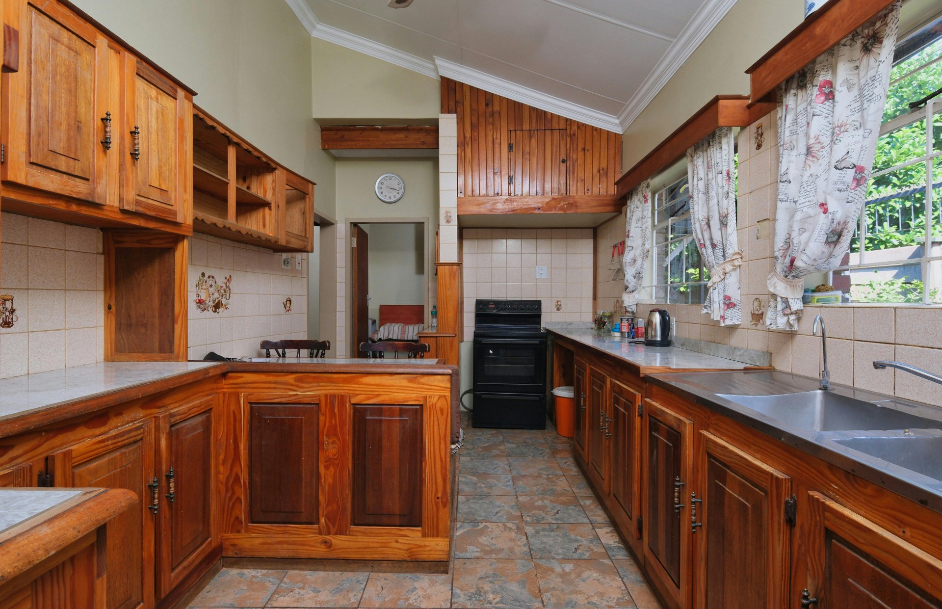 Narrow kitchen with wooden cabinets, black countertops, tiled floor, and a window with curtains and plants.