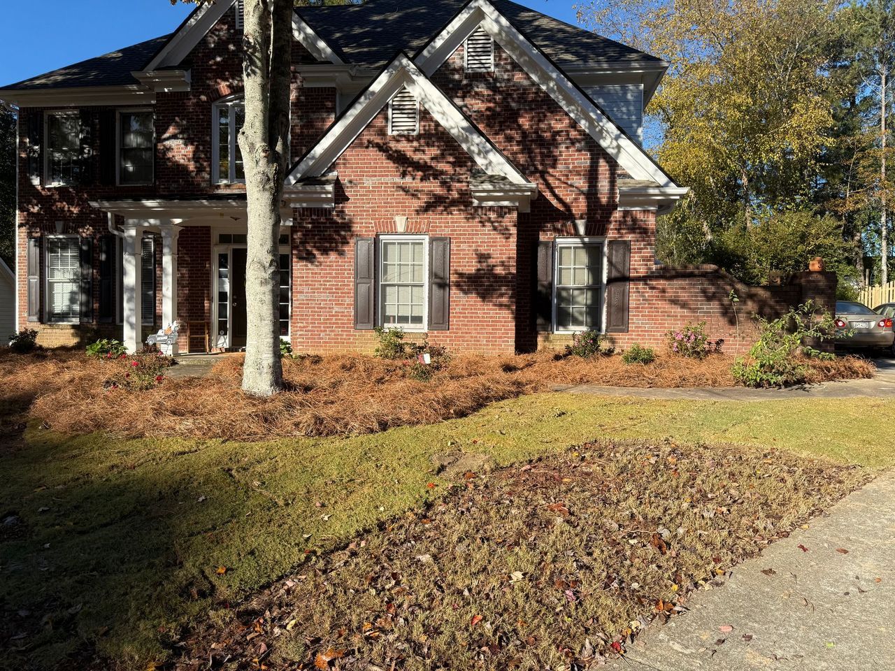 A two-story red brick house with a white trim, front porch columns, and a lawn covered in fallen leaves under a clear sky.