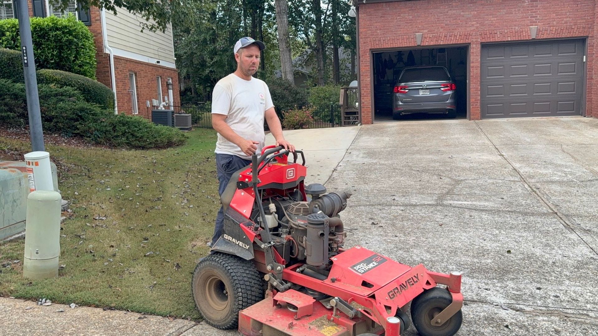 A person in a white shirt and cap stands on a red stand-on commercial lawn mower on a residential driveway.