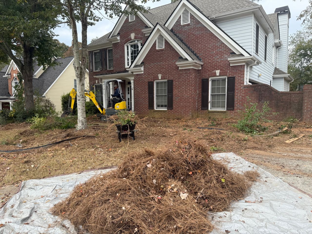 A front yard with a large pile of yard waste on a tarp, with a small excavator parked near the house entrance.