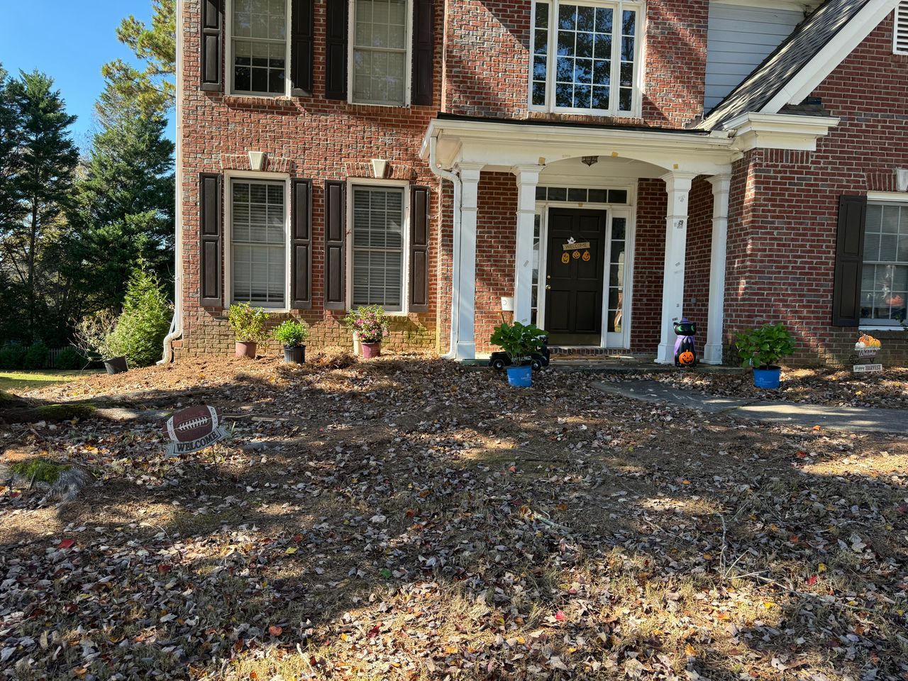 A two-story brick home features a covered white porch entrance surrounded by a yard covered in fallen autumn leaves.