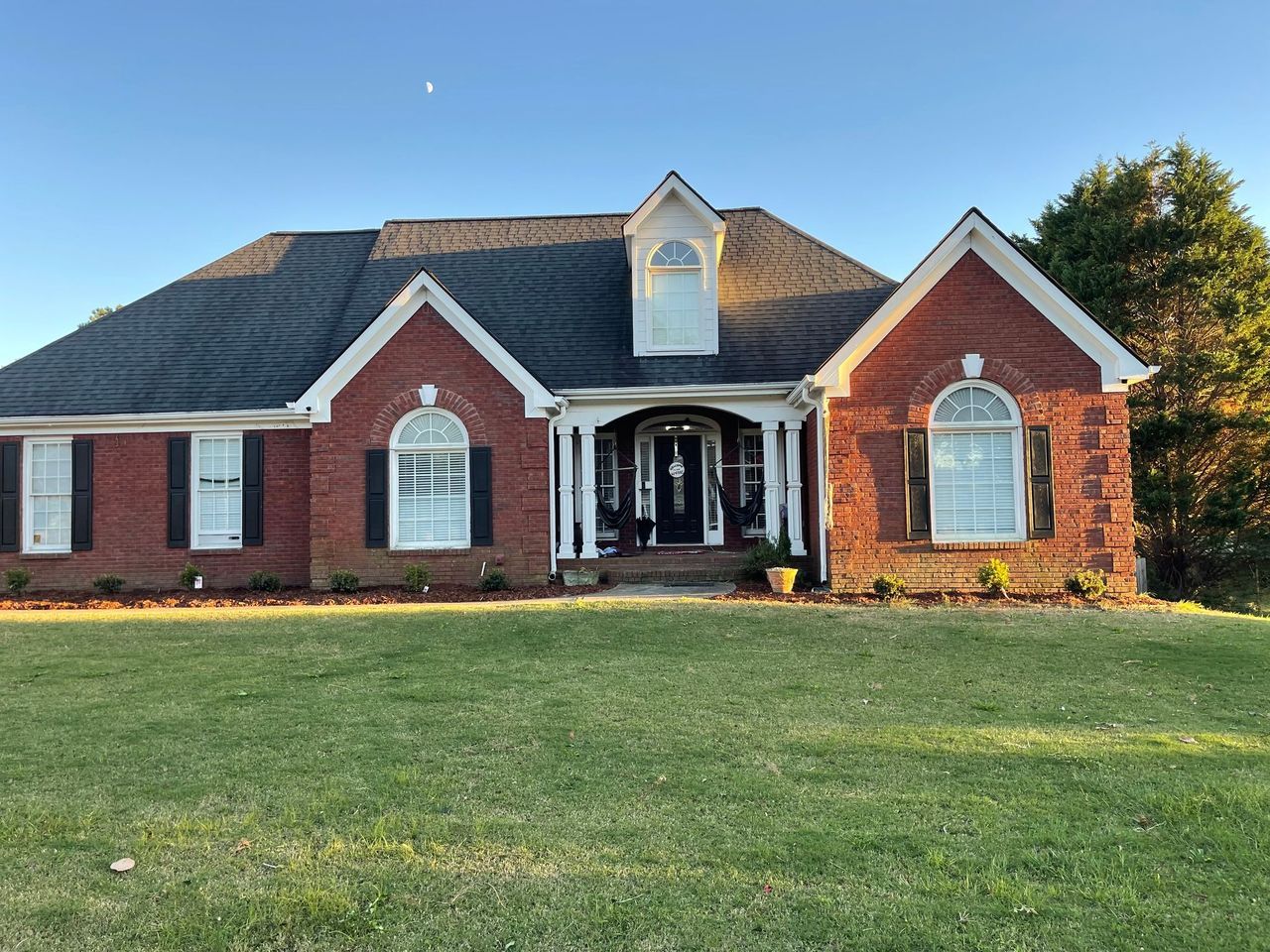 A one-story brick house with a dark shingled roof, arched windows, and white columns at the front entryway on a lawn.