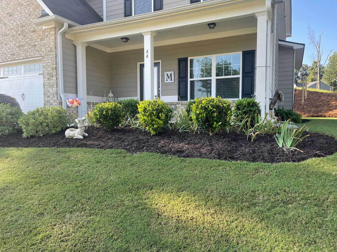 A front yard landscape with a mulched garden bed, green shrubs, a deer statue, and a covered porch on a house.