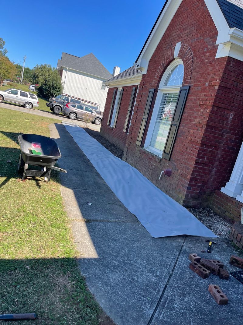 A red brick house exterior with a white plastic tarp laid on the walkway beside a wheelbarrow and scattered bricks.