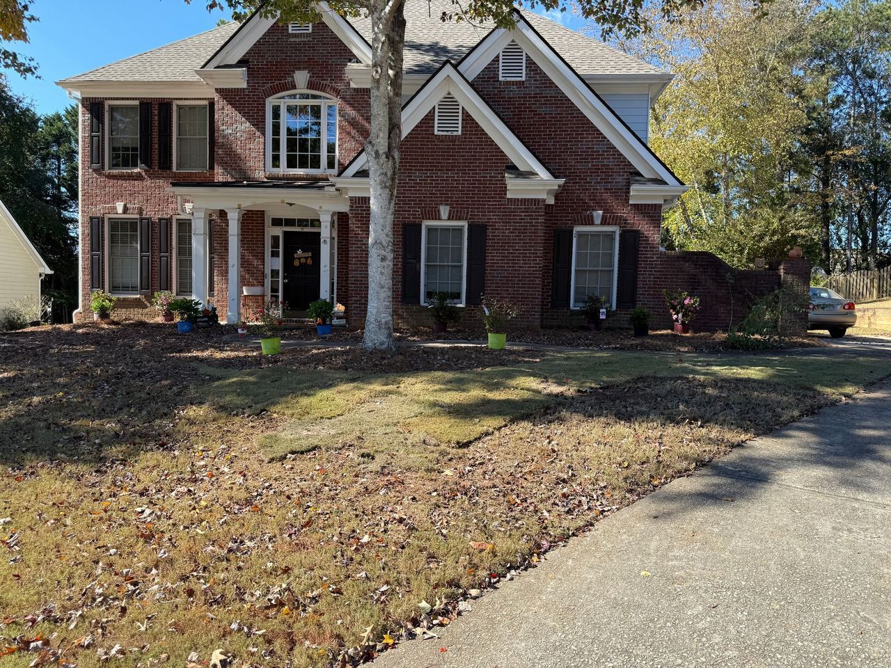 A two-story red brick house with white trim, a covered porch, and a front lawn covered in fallen autumn leaves.