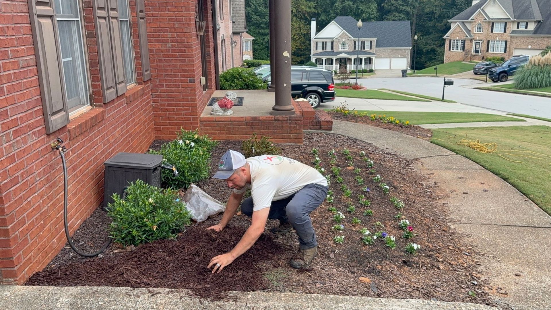 A person wearing a hat and light shirt kneeling to spread mulch in a garden bed in front of a brick house.