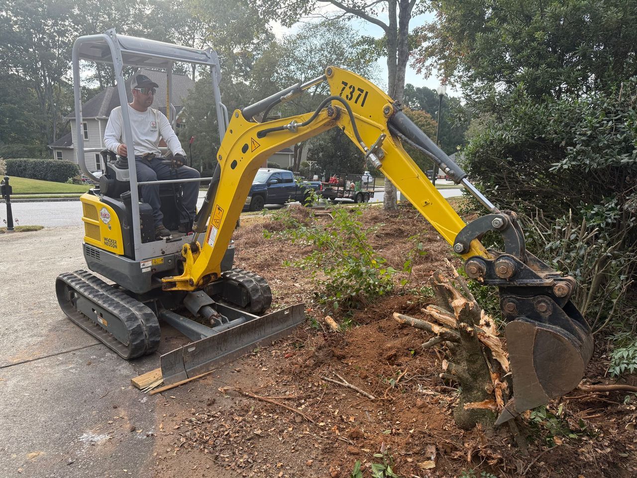 A person operates a yellow compact excavator to remove a tree stump in a residential yard.