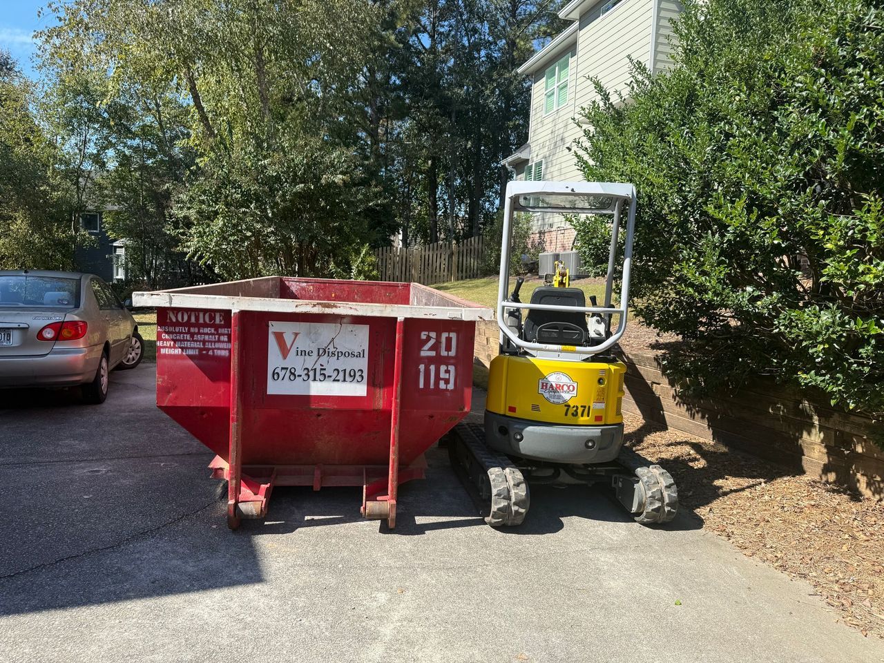 A red dumpster and a small yellow excavator parked on a driveway next to a house and a silver sedan.