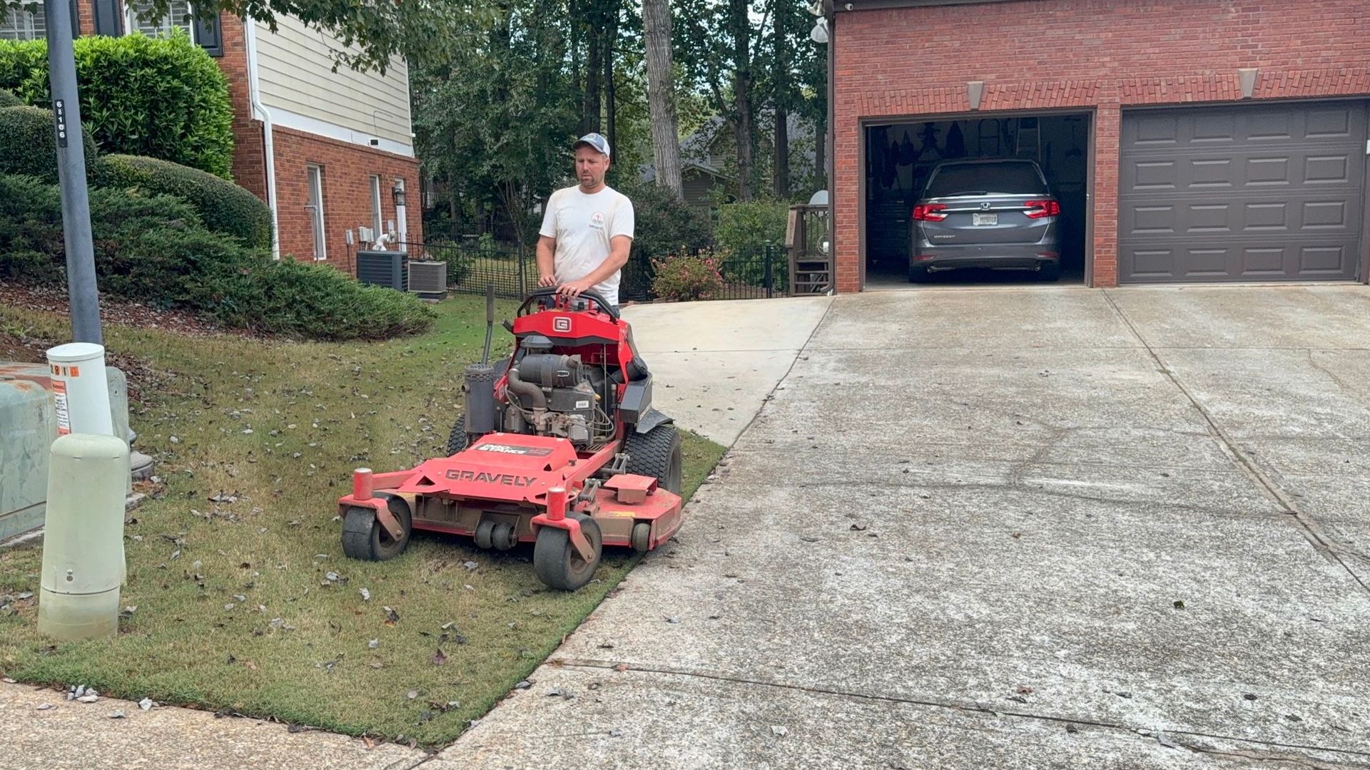 A person drives a red stand-on lawn mower on a residential lawn next to a paved driveway and brick house.
