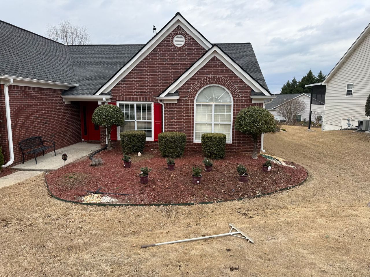 A brick house with a front garden featuring shrubs and red mulch, with a lawn rake lying on the grass in the foreground.