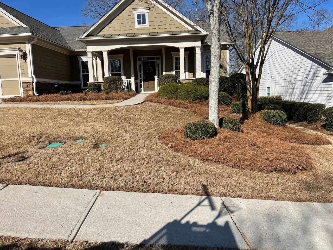 A single-story suburban house with brown siding, a covered porch, and a lawn with dormant grass and mulch beds.