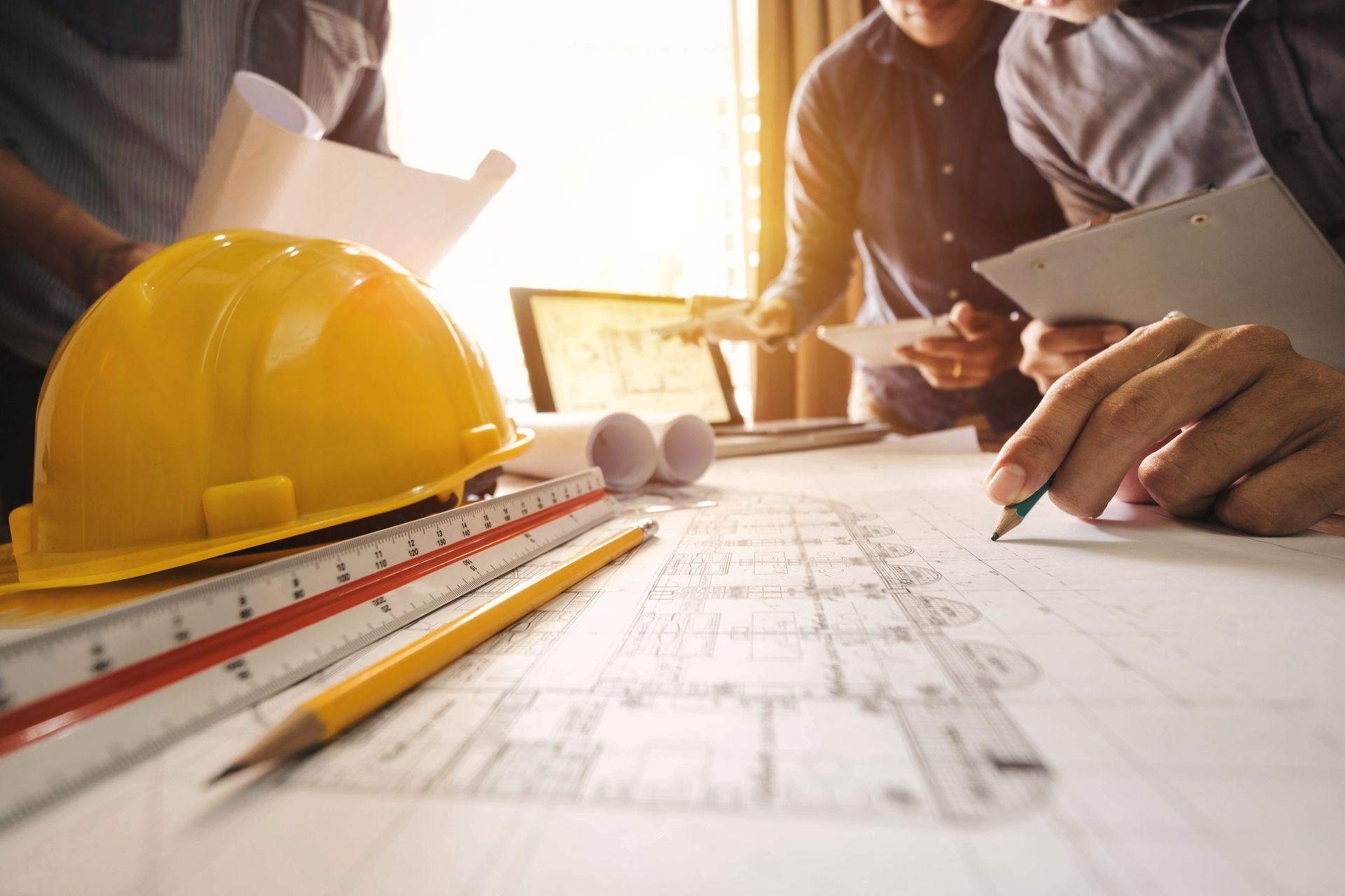 Casque jaune et crayon sur des plans architecturaux, des mains professionnelles examinant les plans.