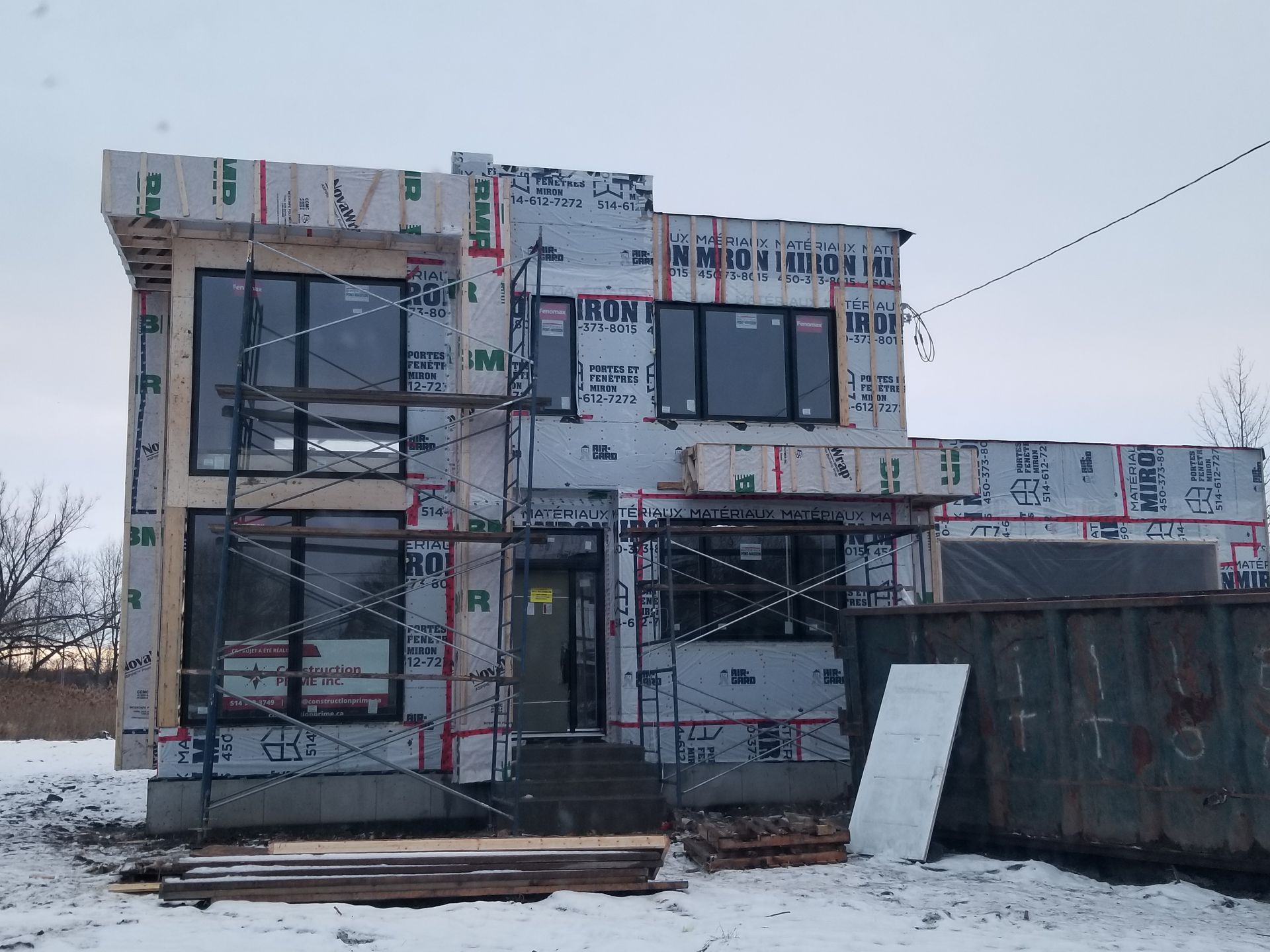 Une maison à deux étages en construction, recouverte d'une bâche blanche résistante aux intempéries, avec des fenêtres sombres, des échafaudages et de la neige.