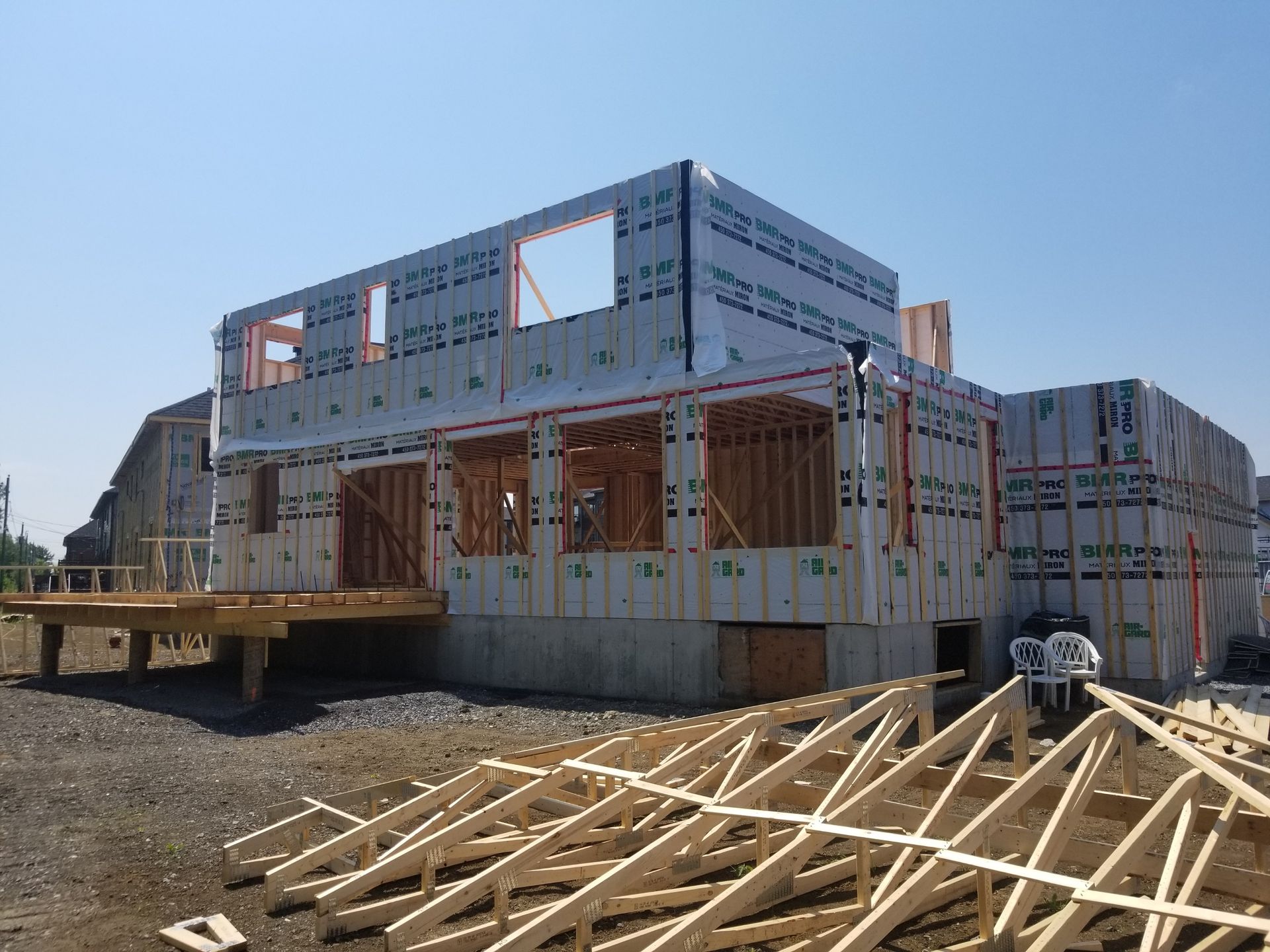 Au premier plan, une maison en construction avec un revêtement extérieur blanc, une terrasse en bois et une charpente de toit en bois.