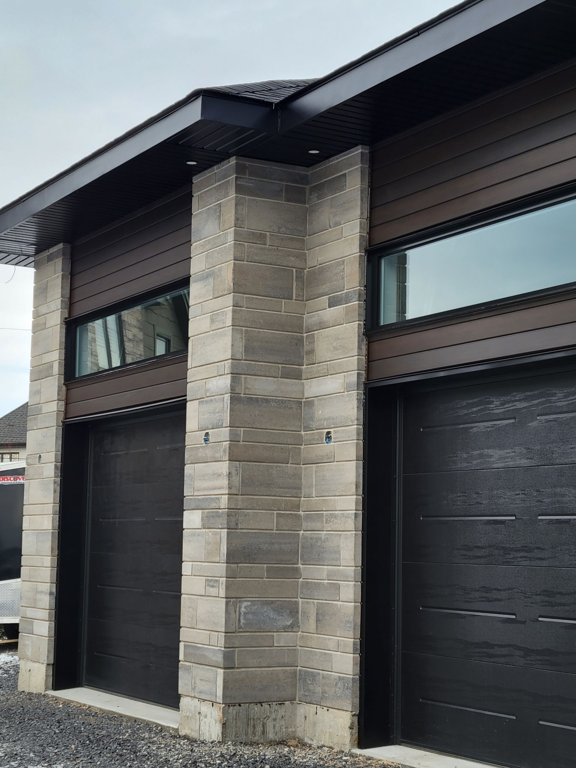 Gros plan sur l'extérieur d'une maison moderne présentant un bardage horizontal foncé, des piliers en pierre et deux portes de garage noires.