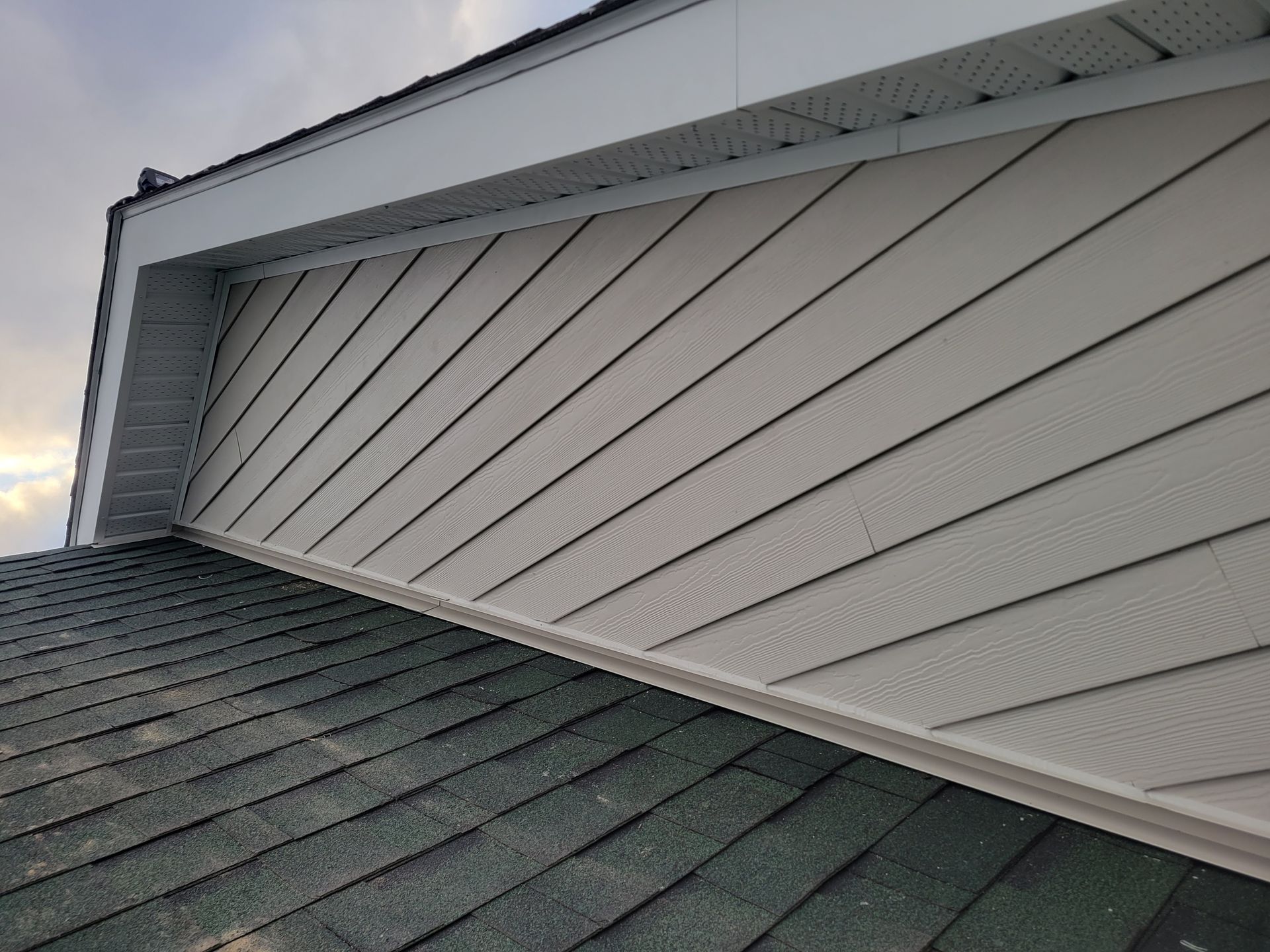 Vue d'un toit recouvert de bardeaux gris foncé et d'un mur à pignon habillé d'un bardage beige clair en diagonale.
