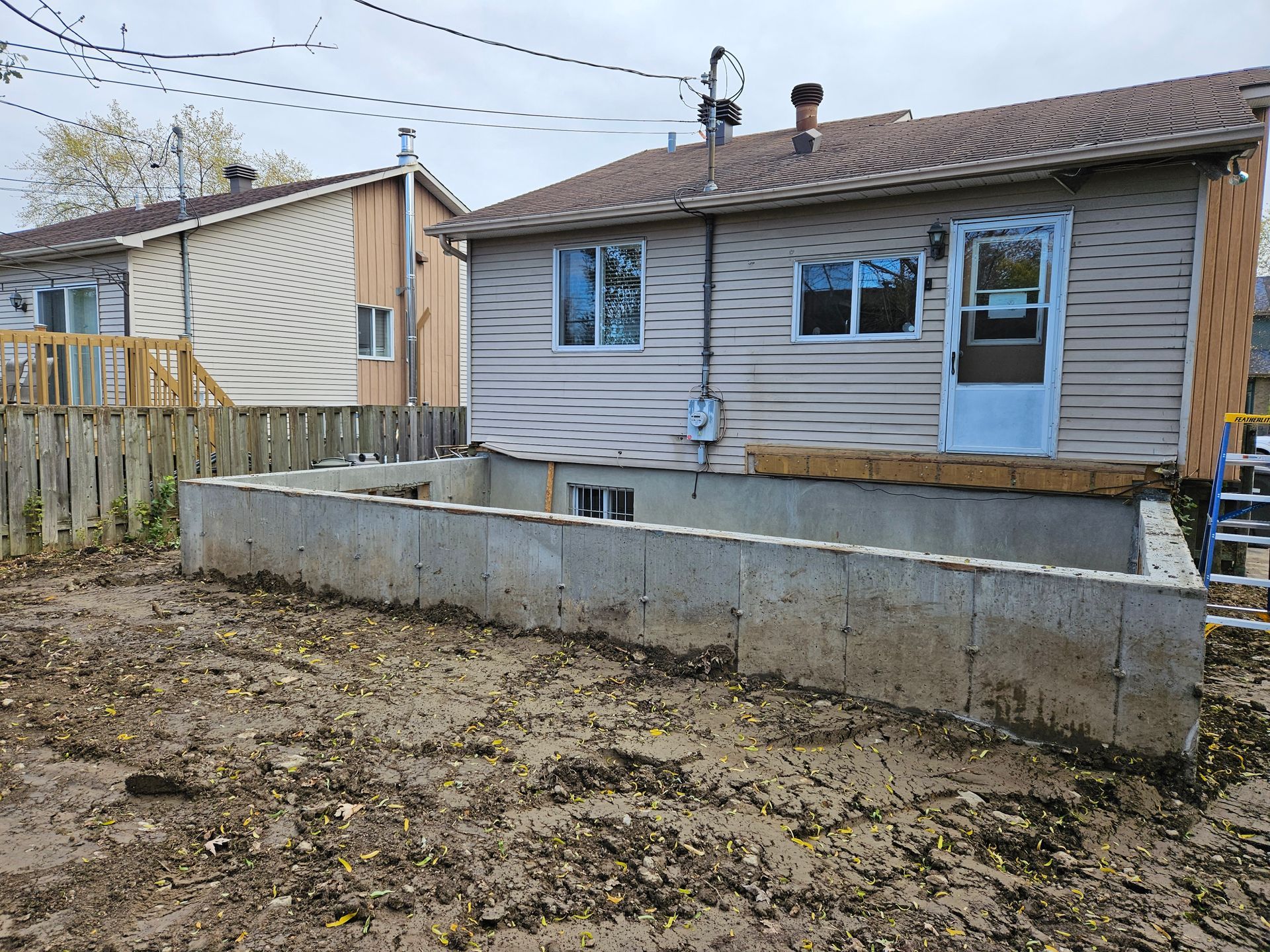 Une cour arrière montrant les fondations en béton fraîchement coulées d'une extension de maison, adjacente à une maison existante.