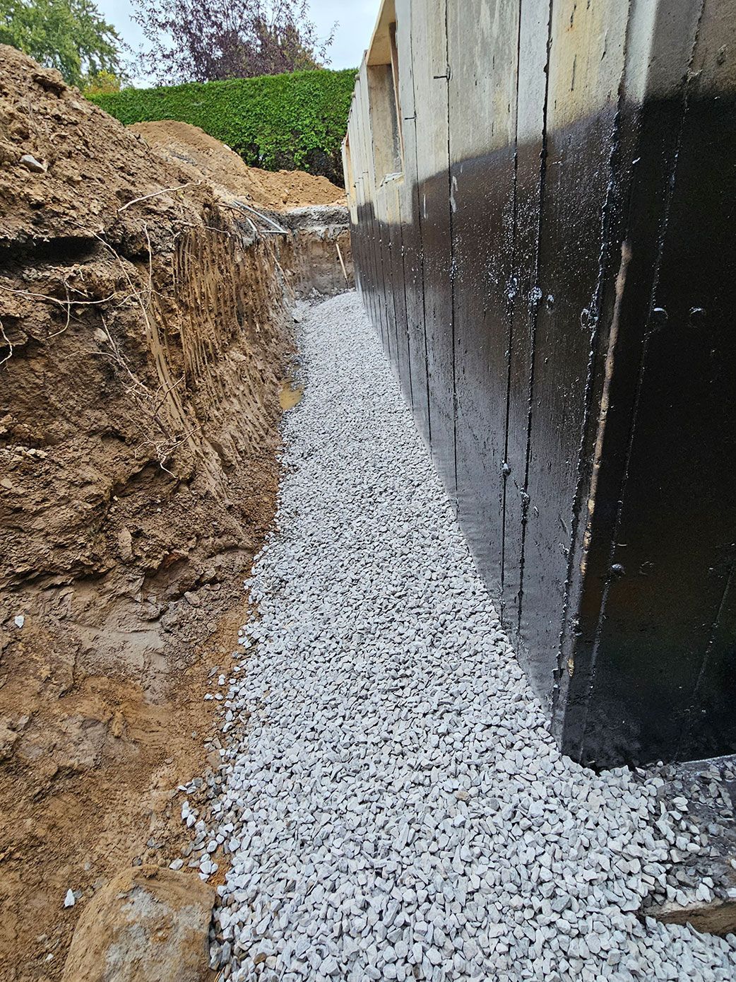 Une tranchée le long d'un mur de fondation, revêtue d'un enduit imperméabilisant noir, remplie de gravier de drainage gris.