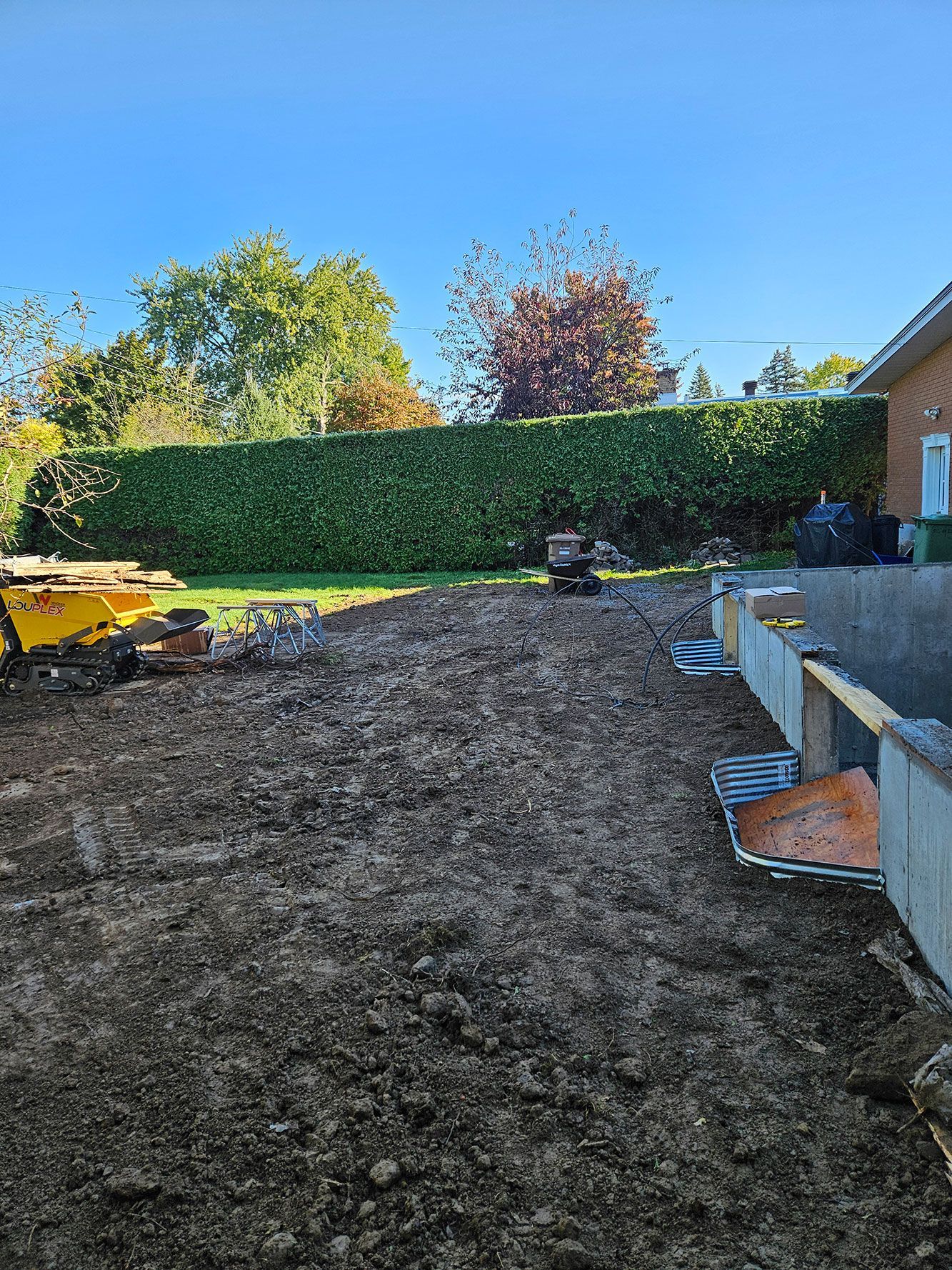 Une cour recouverte de terre, avec une grande haie verte en arrière-plan et des matériaux de construction sur le côté, sous un ciel bleu dégagé.