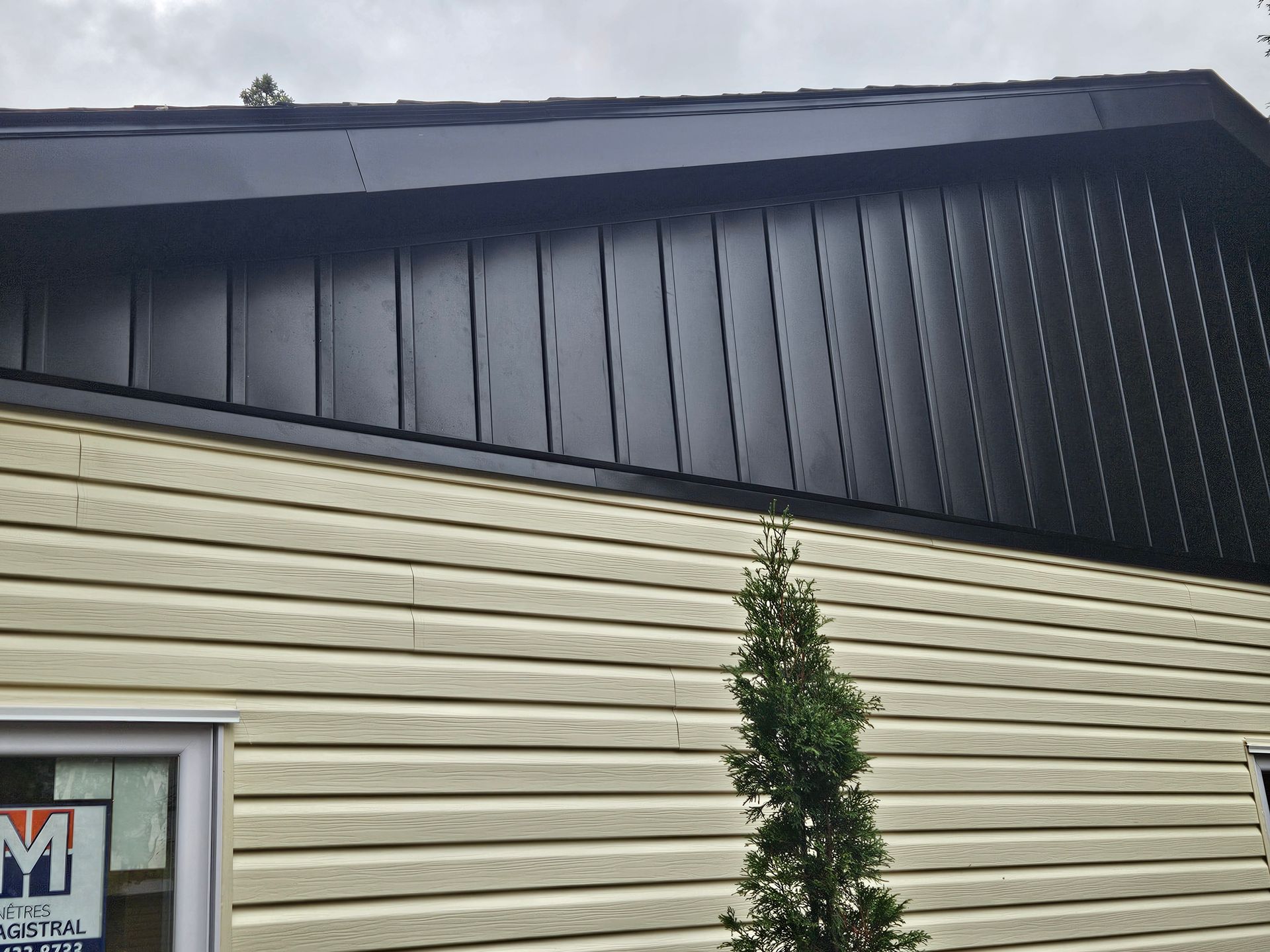 L'extérieur d'une maison présente un revêtement en vinyle jaune clair horizontal sous des panneaux métalliques verticaux noirs sur le pignon du toit.