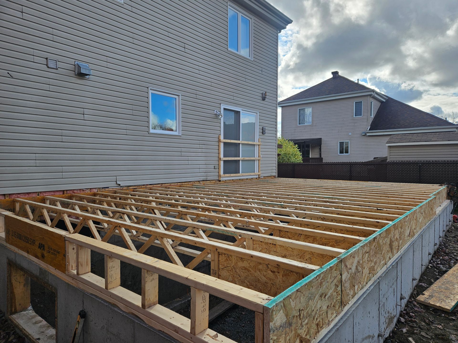 Une structure de terrasse en bois partiellement construite, adossée à l'arrière d'une maison beige à deux étages, sous un ciel nuageux.