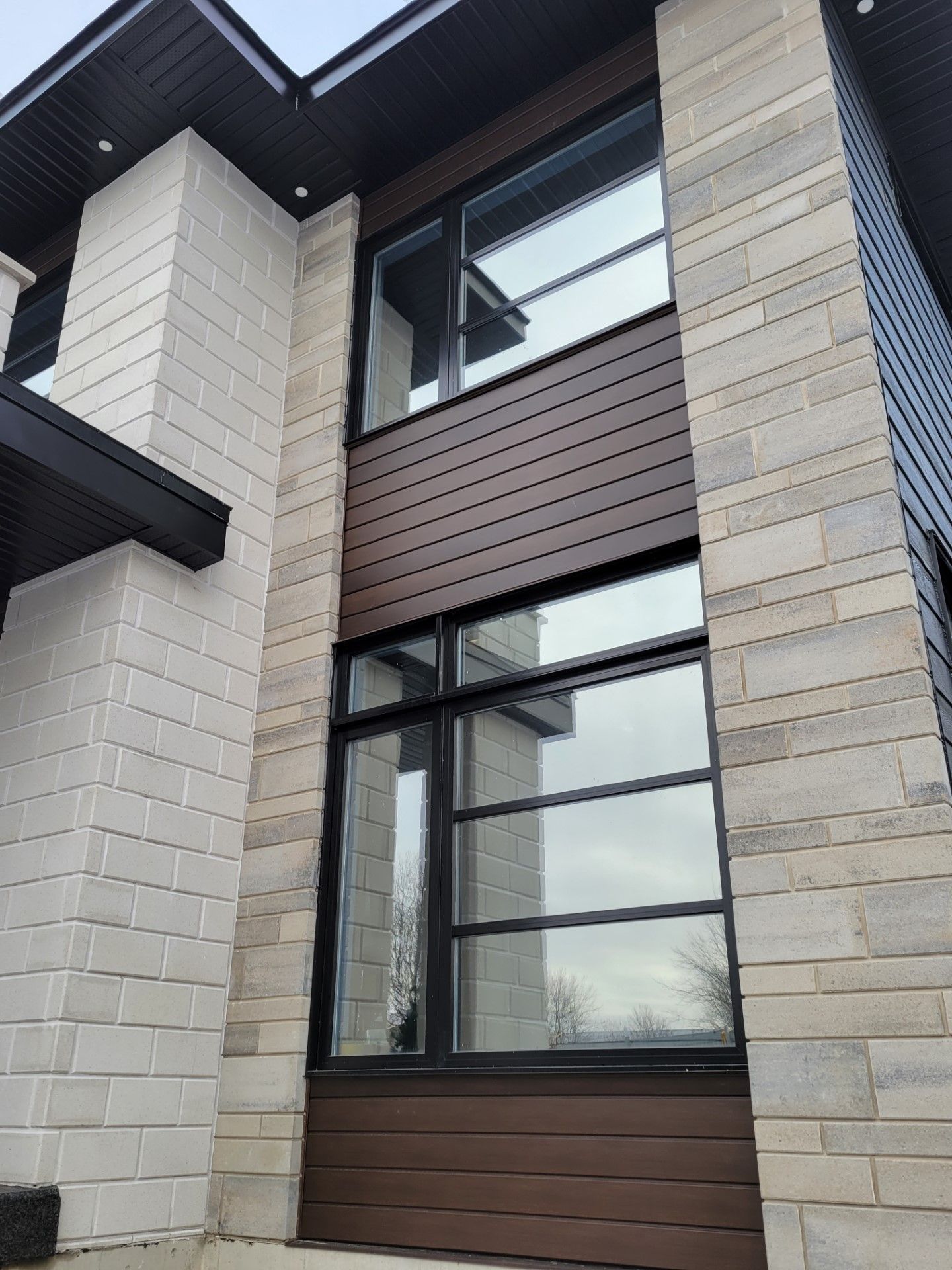Extérieur d'une maison moderne à deux étages, caractérisé par une maçonnerie en pierre claire, un bardage horizontal en bois foncé et de grandes fenêtres rectangulaires.