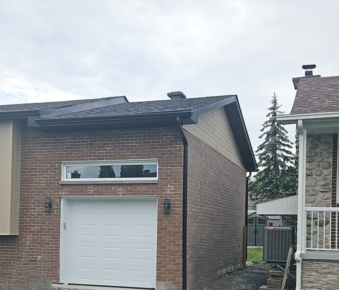 Un garage en briques avec une porte blanche, une fenêtre horizontale au-dessus et un toit en métal sombre, jouxtant une maison.