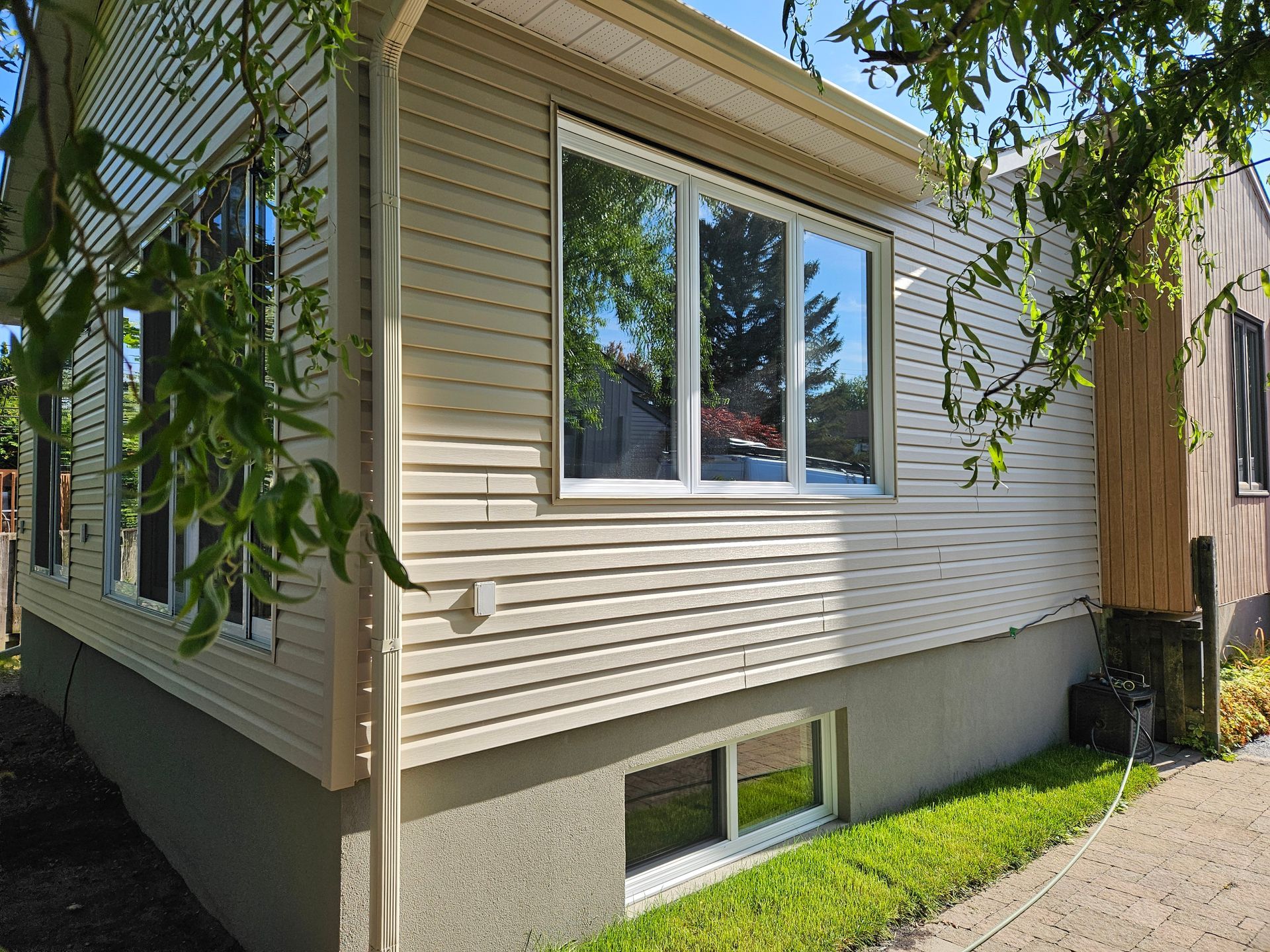 Revêtement extérieur en vinyle beige clair, horizontal, sur le coin extérieur d'une maison avec deux grandes fenêtres et une petite fenêtre au sous-sol.