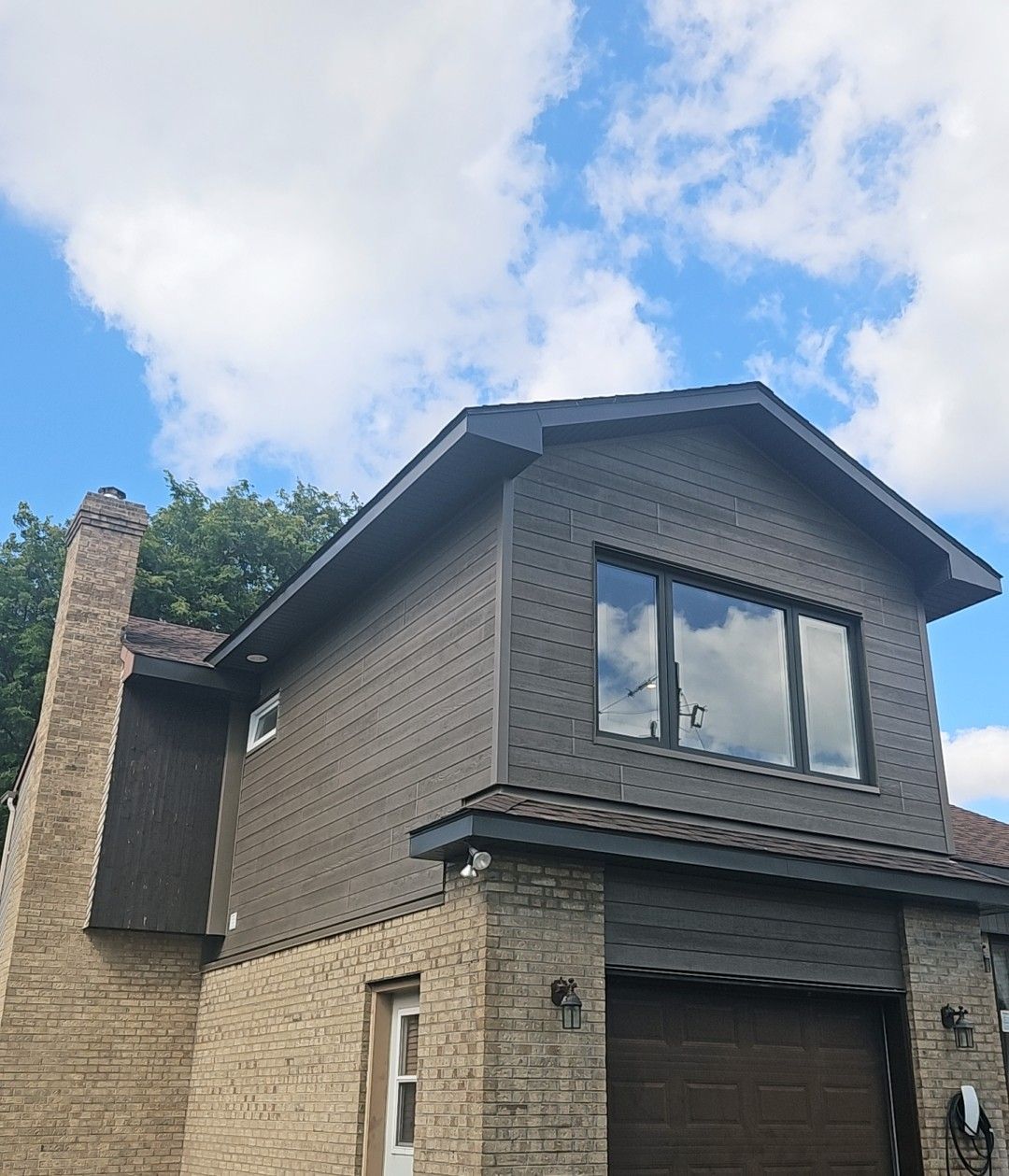 Une maison à deux étages avec un rez-de-chaussée en briques beiges, un étage supérieur gris texturé, une porte de garage foncée et une cheminée en pierre.