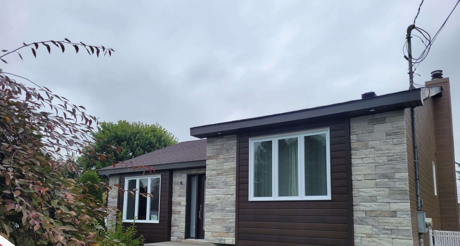 Une maison de plain-pied avec un bardage brun foncé et des accents de pierre gris clair, sous un ciel couvert.
