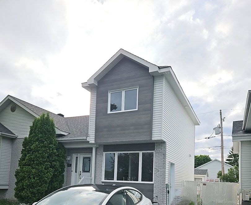 Une maison à deux étages avec un revêtement blanc et des panneaux gris imitation bois, avec une fenêtre en façade et une voiture garée dans l'allée.