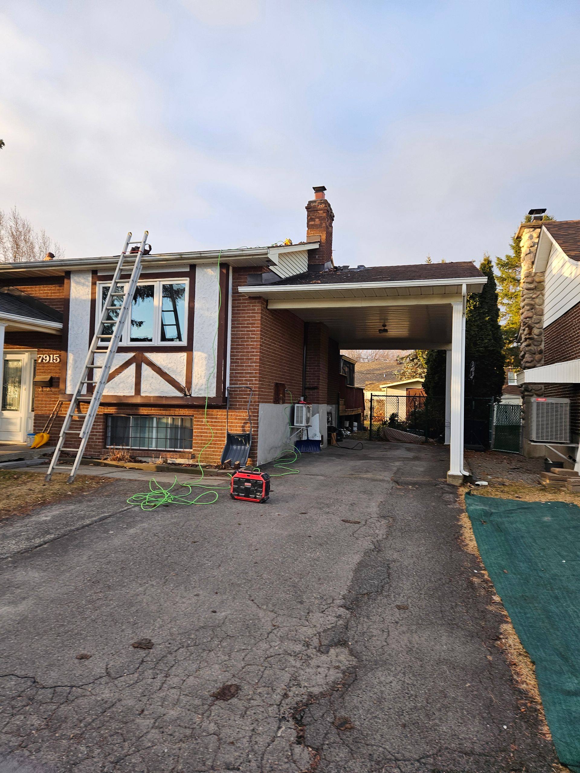 Une maison en briques avec une échelle appuyée contre la façade, en cours de réparation de la toiture, comprenant un abri voiture et une allée asphaltée.
